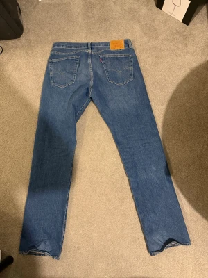 Levi's 501 blå raka jeans - Klassiska blå Levi's 501 jeans med rak passform och femficksmodell. Tillverkade i slitstark denim med ikonisk läderpatch bak i midjan. Perfekta för dig som gillar tidlös stil och vill ha ett par jeans som alltid funkar.