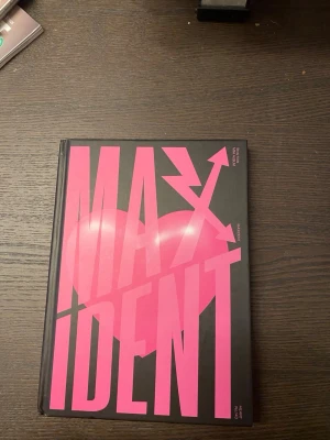 MAXIDENT av Stray Kids - Säljer boken MAXIDENT av Stray Kids. Omslaget är svart med stora rosa bokstäver och ett hjärta i bakgrunden. Boken har hårda pärmar och en modern, grafisk design. Perfekt för dig som gillar K-pop och vill ha något unikt i bokhyllan.