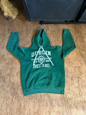 Grön Dublin Ireland hoodie - Snygg grön hoodie med vit text 'Dublin Ireland' och tryck framtill. Har stor huva med vita snören och en klassisk magficka. Perfekt för dig som gillar streetwear och vill ha något bekvämt och stilrent. Materialet känns mjukt och skönt mot huden.