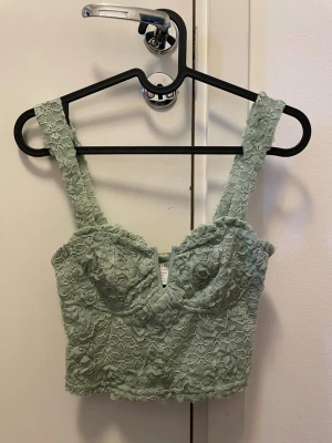 Grön spets korsettopp - Snygg ljusgrön korsettopp med breda axelband och v-ringad urringning. Toppen är täckt av blommig spets och har en croppad passform som ger en trendig look. Perfekt att styla med höga jeans eller kjol för en cool outfit.