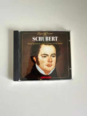 CD: Franz Schubert String Quartets  - Klassisk CD med Franz Schuberts stråkkvartetter nr 13 och 14, inklusive 'Death and the maiden'. Omslaget är svart och gult med porträtt och gul text. Plastfodral med tryckt baksida där låtar och speltid står. Perfekt för dig som gillar klassisk musik.
