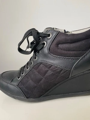 Svarta kilklackade sneakers - Snygga svarta sneakers med kilklack, quiltade detaljer på sidorna och kombination av mocka och läder. Skorna har svarta snören, dragkedja på sidan och högt skaft för extra support. Perfekta för dig som vill ha både stil och komfort.