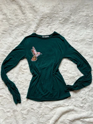 Mörkgrön topp med fågelapplikation - Mörkgrön långärmad topp med en stor broderad fågelapplikation på bröstet. Toppen har rund halsringning och lätt rynkade ärmar för en cool detalj. Perfekt för dig som vill ha något unikt och stilrent i garderoben.