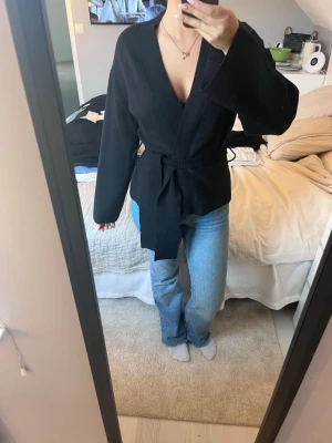 Svart omlottkofta med knytband - Snygg svart omlottkofta med brett knytband i midjan och vida ärmar. Koftan har en djup V-ringning och är perfekt att styla över ett linne eller t-shirt. Passar till både jeans och kjol för en trendig look.