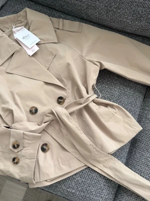Kort beige trenchcoat med bälte - Stilren kort trenchcoat från Gina Tricot. Slutsåld online och dem flesta butiker.