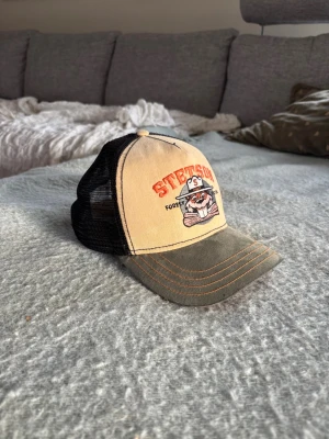 Beige och grön Stetson truckerkeps - Säljer en beige och grön truckerkeps från Stetson med svart mesh på sidorna och baksidan. Framtill finns ett broderat motiv av en björn och texten 'STETSON FOREST'. Kepsen har böjd skärm och justerbar snapback. Perfekt för en avslappnad streetwear-look.