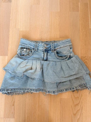 Kort jeanskjol med volanger - Säljer en ljusblå jeanskjol med dubbla volanglager och råa kanter. Kjolen har bälteshällor och bakfickor för en cool och avslappnad look. Perfekt för dig som gillar denim och vill ha en trendig kortkjol i garderoben.