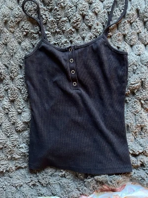 Svart ribbad linne med knappar - Snyggt svart ribbat linne med smala axelband och fyra dekorativa metallknappar framtill. Perfekt att styla till jeans eller kjol för en trendig look. Materialet är stretchigt och följsamt.