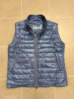 Blå dunväst från Herno, stl L/52 - Snygg blå dunväst från Herno med quiltad design och hög krage. Västen har två dragkedjeförsedda fickor framtill och stängs med dragkedja. Tillverkad i lätt, glansigt material som ger en modern look. Perfekt för lager-på-lager-stil.