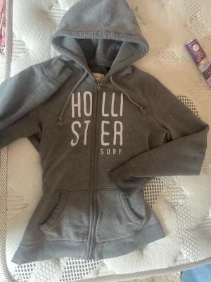 Hollister hoodie - Säljer en grå hoodie från hollister med dragkedja och vit text på bröstet. Hoodien har huva med snören, två fickor framtill. Det ör storlek L men jag har sytt den tajtare som ni ser på bilden skulle säga att den är xs-s super skön. Säljer för den inye kommer till användning 💗💗