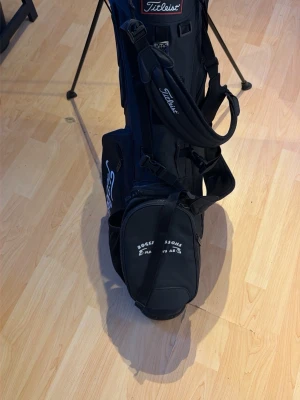 Svart golfbag från Titleist - Säljer en svart golfbag från Titleist med vita logotyper och flera praktiska fack. Väskan har axelremmar, bärhandtag och stödben för att stå stadigt på marken. Perfekt för att organisera och bära golfklubbor och tillbehör på banan.