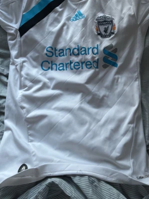 Liverpool FC vit matchtröja Adidas - Säljer en vit Liverpool FC matchtröja från Adidas med klubbmärke, sponsorlogga och blå detaljer. Tröjan har rund hals, korta ärmar och är tillverkad i ett lätt funktionsmaterial som andas. Perfekt för fotbollsträning eller att bära på läktaren. Har en liten fläck som går bort i tvätten 