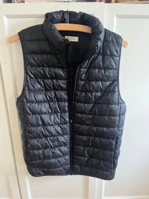 Svart dunväst från H&M - Snygg svart dunväst från H&M med quiltad design och hög krage. Västen har dragkedja framtill och två sidofickor. Perfekt för lager på lager och ger en clean look till din outfit. Tillverkad i lätt och värmande material.
