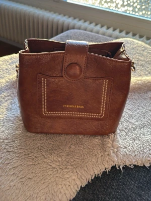 Brun axelväska Fashion & Bags - Nästan oanvänd. Snygg brun axelväska i konstläder från Fashion & Bags. Väskan har en fyrkantig form, dekorativ söm på framsidan och ett brett, mönstrat axelband med guldfärgade detaljer. Praktisk ficka framtill och stängs med tryckknapp. Mått 20x20x10