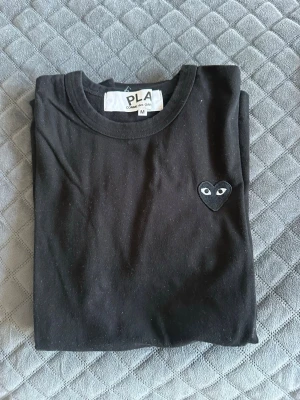 Svart t-shirt från Comme des Garçons - Säljer en svart t-shirt från Comme des Garçons Play med det ikoniska hjärta med ögon broderat på bröstet. Klassisk rund hals och normal passform. Perfekt basplagg med en stilren twist. Perfekt inför sommaren och ett väldigt bra skick. Skriv om du vill ha mer info eller fler bilder. Kan inte vissa den på för den är för liten för mig!!!