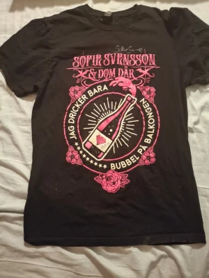 Svart Gildan t-shirt med rosa tryck - Cool svart t-shirt från Gildan med stort rosa tryck på bröstet. Motivet har texten 'Sofie Svensson & Dom Där' och 'Jag dricker bara bubbel på balkongen' samt en flaska och rosor. Klassisk rund hals och korta ärmar. Perfekt för dig som gillar statement-plagg.