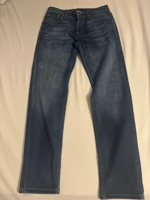 Blå raka jeans med slitningar - Säljer ett par klassiska blå jeans med raka ben och diskreta slitningar framtill. Jeansen har fem fickor, normal midja och är tillverkade i ett mjukt denimtyg. Perfekta för en avslappnad och stilren look.