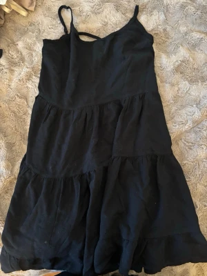 Svart sommarklänning med volanger - Svart kortklänning med tunna axelband och volangdetaljer. Klänningen har en luftig passform och är tillverkad i ett mjukt tyg, perfekt för varma dagar. Dragkedja i sidan för enkel påklädning. Enkel och stilren design som passar till många tillfällen.