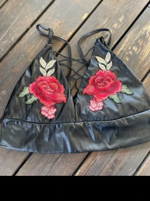 Svart bralette med rosbrodyr från LANSHTER - Trendig svart bralette i konstläder från LANSHTER med broderade röda rosor och gröna blad på kuporna. Axelbanden är smala och korsas i ryggen för en snygg detalj. Perfekt för dig som vill ha något unikt och edgy i garderoben.