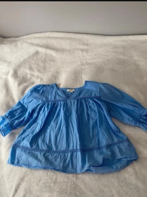 Blå vid blus med lång ärm - Blå luftig blus med v-ringning och långa ärmar. Blusen har en avslappnad passform med rynkade detaljer och dekorativa sömmar. Tillverkad i ett lätt bomullsliknande material som känns skönt mot huden. Perfekt för en avslappnad och trendig look. Liten defekt vid armhålorna men inget som stör hela looken.(kolla bild)