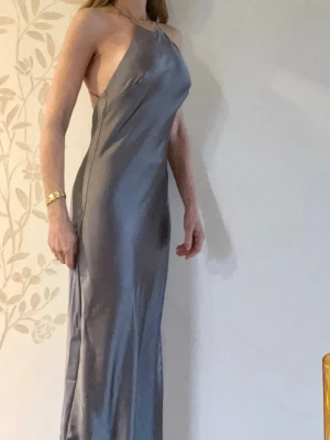 Silverfärgad långklänning i satin - Snygg blå/silverfärgad långklänning i satin, stl M med halterneck och öppen rygg. Klänningen har en slimmad passform och tunna axelband som korsas i ryggen. Jag är xs, så det sitter alldeles för löst på mig på bilden.