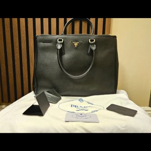 Prada väska Saffiano-läder - Elegant Prada väska i svart Saffiano-läder i mycket fint skick. Klassisk och tidlös modell som passar både till arbete och vardag. Väskan är rymlig och håller formen fint. Prada dustbag och autenticitetkort medföljer (se bilder). Väl omhändertagen och sparsamt använd. Säljs då den inte används tillräckligt. Kan skickas direkt vid snabb affär.