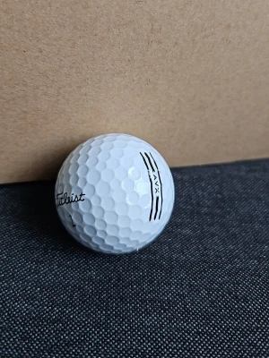 Vit golfboll Titleist AVX - Säljer en vit golfboll av modellen AVX från Titleist. Bollen har klassiskt dimple-mönster och svarta tryckta linjer samt logga. Perfekt för dig som gillar golf och vill ha en boll med bra känsla och kontroll.