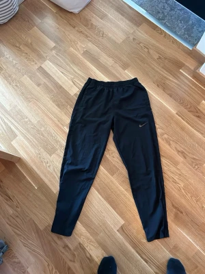 Svarta Nike Dri-FIT träningsbyxor - Säljer ett par svarta träningsbyxor från Nike med Dri-FIT-material som transporterar bort fukt. Byxorna har en smal passform, elastisk midja och diskret Nike-logga på benet. Perfekta för gymmet eller löpning.