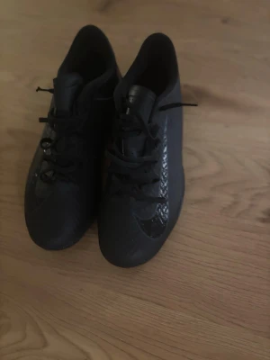 Svarta Nike fotbollsskor - Säljer ett par helsvarta fotbollsskor från Nike med låg profil och snörning. Skorna har en modern design med blanka detaljer på sidorna och är tillverkade i syntetmaterial för bra grepp och kontroll på planen. Perfekta för dig som vill ha stilrena och funktionella skor.