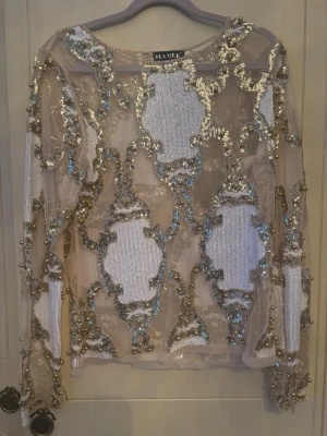 Glittrig topp med paljetter  - Säljer en transparent glittrig topp med långa ärmar. Toppen är dekorerad med guld- och silverfärgade paljetter och pärlor i ett unikt mönster. Perfekt för dig som vill sticka ut med en statement piece.