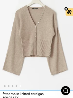 Beige stickad cardigan med vida ärmar - Trendig beige stickad cardigan från ginat med v-ringning, knappar framtill och bred ribbstickad midja. Modellen har vida ärmar och croppad passform som ger en modern siluett. Perfekt att styla med jeans eller kjol för en avslappnad look.