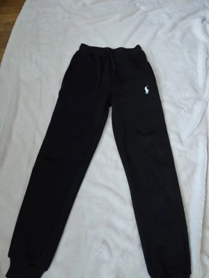 Svarta mjukisbyxor från Polo Ralph Lauren - Svarta mjukisbyxor från Polo Ralph Lauren med klassisk vit logga på vänster höft. Byxorna har dragsko i midjan och ribbade muddar vid bensluten. Perfekta för chill dagar och har en skön passform i mjukt material.