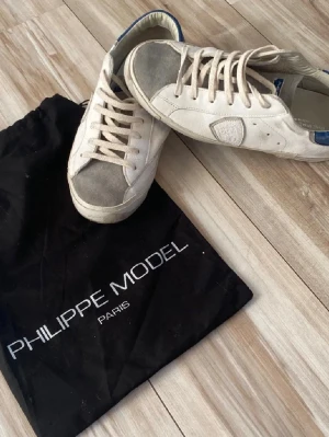 Philippe Model  - Snygga vita sneakers från Philippe Model med grå mockadetaljer på tån och blå detaljer vid hälen. Klassisk låg modell med vita snören och gummisula där det står 'PHILIPPE MODEL' undertill. Perfekta för en clean och trendig look i sommar. Hör av er för om ni undrar något. Pris kan diskuteras.