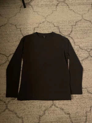 Svart långärmad tröja från SMOG - En enkel och stilren svart långärmad tröja från SMOG med relaxed fit. Tröjan har rund halsringning och är tillverkad i ett mjukt, strukturerat material som ger en avslappnad look. Perfekt att matcha med jeans eller chinos.