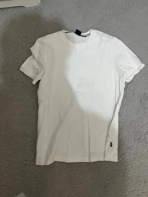 Vit basic t-shirt från Hugo boss - Säljer en klassisk vit t-shirt från Hugo boss med rund hals och korta ärmar. T-shirten har en diskret logga på bröstet och är tillverkad i mjuk bomull. Perfekt till jeans eller shorts för en clean look.