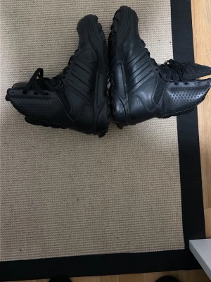 Svarta taktiska kängor i skinn - Säljer ett par svarta taktiska kängor med robust sula och snörning hela vägen upp. Tillverkade i slitstarkt skinn och syntetmaterial, perfekta för dig som vill ha en cool och funktionell look. Kängorna har förstärkta partier och sportig design. Vatten täta