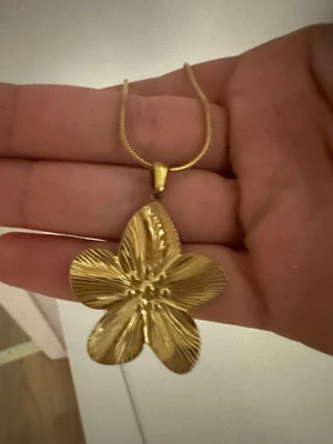 Guldigt halsband med blomma - Snyggt halsband med ett stort, detaljerat blomformat hänge i guldfärg. Kedjan är tunn och elegant, och blomman har en skimrande yta med fina linjer som ger ett unikt intryck. Perfekt för dig som vill ha ett statement-smycke med naturinspirerad design.