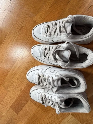 Vita Nike Air Force 1 sneakers - Klassiska vita Nike Air Force 1 sneakers med lågt skaft och perforerad tåbox. Skorna har en chunky sula, vita skosnören och diskreta Nike-loggor på sidan. Tillverkade i läder med meshfoder för extra komfort. Perfekta för en clean och stilren look.