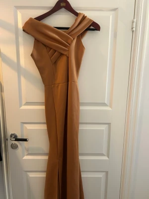 Orange/Beige långklänning med omlott-detalj - Snygg orange/beige långklänning med unik omlott-detalj över axeln och asymmetrisk skärning. Klänningen har en elegant och figurnära passform, perfekt för speciella tillfällen. Materialet känns mjukt och följsamt.