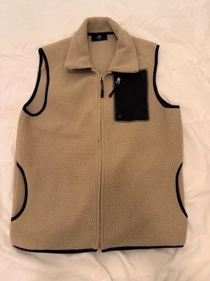 Beige fleeceväst med svart detalj - Beige fleeceväst från H&M med svart bröstficka med dragkedja och svarta kantband. Västen har hel dragkedja framtill och två sidofickor. Perfekt för lager-på-lager och chill stil.