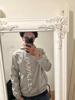 Beige vindjacka från Moncler - Snygg beige vindjacka från Moncler med stor logga längs dragkedjan och klassisk Moncler-patch på ärmen. Jackan har huva, elastiska muddar och dragkedja framtill. Perfekt för dig som vill ha en stilren och trendig look.