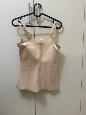 Beige shapewear topp med breda axelband - Shapewear topp i beige med breda, justerbara axelband och formande sömmar. Materialet är slätt och stretchigt för att ge en smickrande siluett. Toppen har hakknäppning bak för enkel på- och avtagning. Perfekt under tighta plagg för en jämn look.