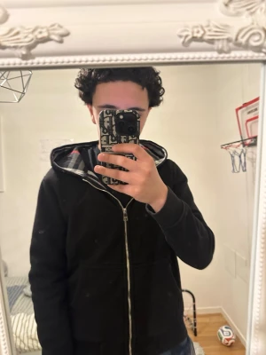 Svart hoodie med rutigt foder - Säljer en svart hoodie med dragkedja och huva. Insidan är fodrad med ett rutigt mönster i svart, vitt och rött. Hoodien har fickor framtill och muddar vid ärmslut och nederkant. Perfekt för dig som gillar en enkel men snygg stil.