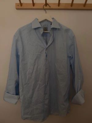 Ljusblå randig skjorta från La chemise  - Klassisk ljusblå skjorta från La chemise  med tunna vita ränder. Skjortan har lång ärm, knappar framtill och en stilren krage. Tillverkad i bomull för en skön känsla och snygg passform. Perfekt för dig som gillar en clean och tidlös stil.