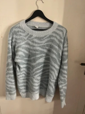 Ljusblå zebramönstrad stickad tröja - Mysig stickad tröja i ljusblått med zebramönster i vitt. Tröjan har rund halsringning, långa ärmar och är fluffig i materialet. Perfekt för dig som gillar djurmönster och vill ha en trendig look. Super skön och sitter fint på! 