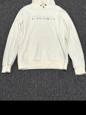 Vit hoodie från Prada med logga - Snygg vit hoodie från Prada med stor ficka framtill och klassisk huva. PRADA-loggan är broderad i rött och svart på bröstet. Hoodien har ribbade muddar vid ärmar och midja, och är gjord i mjukt bomullsmaterial för en skön känsla.