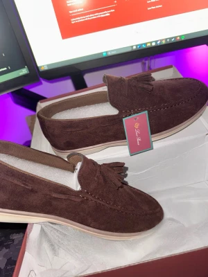 Bruna mocka loafers med tofsar - Snygga bruna loafers i mjuk mocka med dekorativa tofsar framtill. Skorna har en klassisk siluett, ljus sula och diskreta sömmar längs ovandelen. Perfekta för dig som gillar stilrena och bekväma skor med en twist.