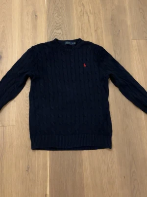 Mörkblå kabelstickad tröja Polo Ralph Lauren - Säljer en mörkblå kabelstickad tröja från Polo Ralph Lauren med klassisk röd logobrodyr på bröstet. Tröjan har rund hals, långa ärmar och ribbade muddar. Perfekt för dig som gillar stilrena och tidlösa plagg.