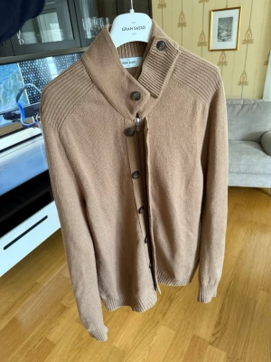 Gran sasso zip - Stilren beige kofta från Gran Sasso med hög krage och knäppning framtill. Koftan har ribbade detaljer vid axlar och krage samt muddar vid ärmslut och nederkant. Perfekt för lager på lager och en snygg vardagslook.