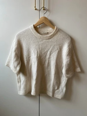 Vit stickad t-shirt i ullblandning - Kortärmad stickad tröja i mjuk ullblandning. Färgen är krämvit och modellen har ribbade kanter vid hals, ärmslut och nederkant. Perfekt för lager på lager och enkel att styla till olika outfits. bra skick inga täcken på slitage 